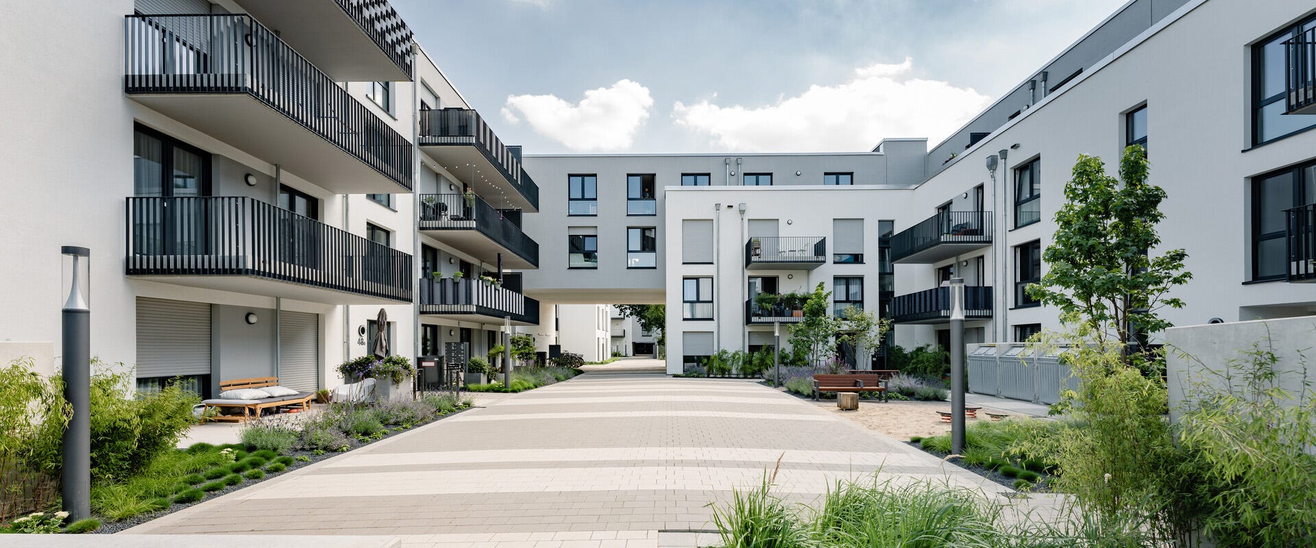 Moderne Wohnanlage mit mehreren weißen Gebäuden, großen Fenstern und dunklen Balkongeländern. Eine begrünte und gepflasterte Fußgängerzone führt durch den Innenhof, gesäumt von Pflanzen, Sitzbänken und stilvollen Straßenlaternen. Die Architektur kombiniert klare Linien mit offenen, einladenden Außenbereichen.