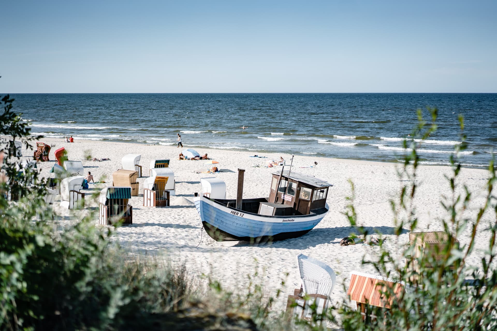Ostseestrand von Heringsdorf mit Strandkörben und einem alten Heringsdorfer Fischerboot, mit Blick auf die Ostsee unter blauem Himmel. Einige Menschen entspannen am Strand oder gehen am Wasser entlang.