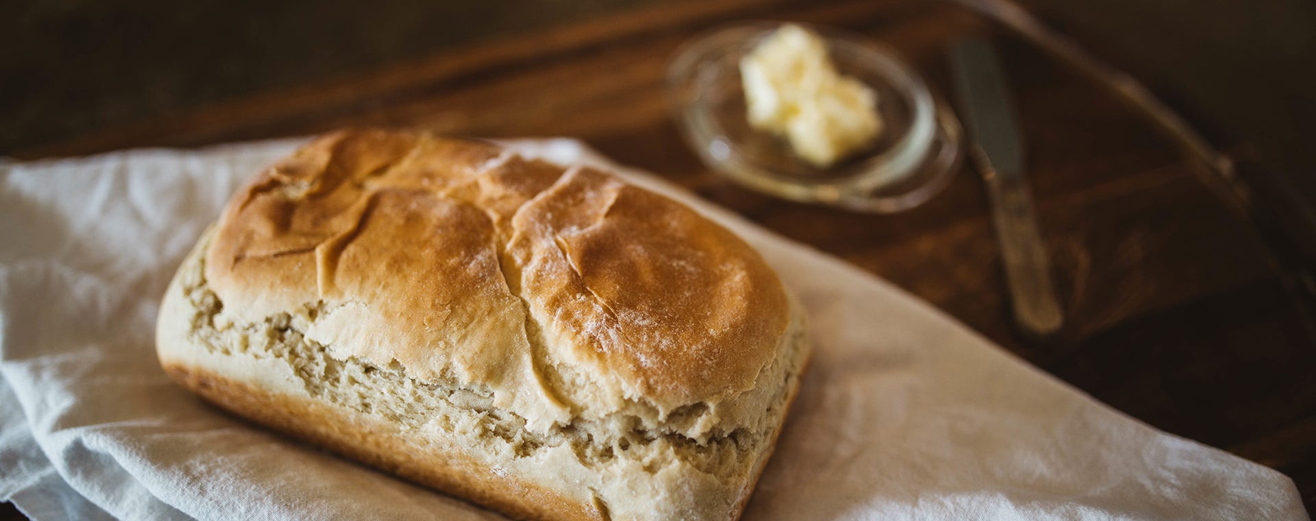 Frisch gebackenes Brotlaib auf einem weißen Tuch; im Hintergrund stehen ein Teller mit Butter und ein Messer auf einem Holztisch.