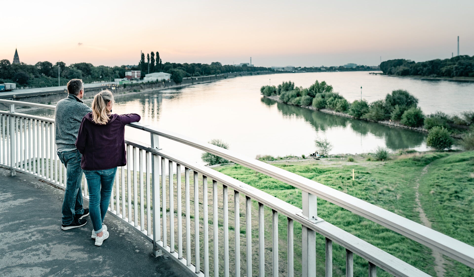 Ein Paar steht auf einer Brücke und blickt auf den ruhigen Fluss in Weidenpesch bei Sonnenuntergang. Die grüne Uferlandschaft und die friedliche Atmosphäre symbolisieren Lebensqualität und Naherholung – ideal für alle, die eine Wohnung kaufen in Weidenpesch möchten.
