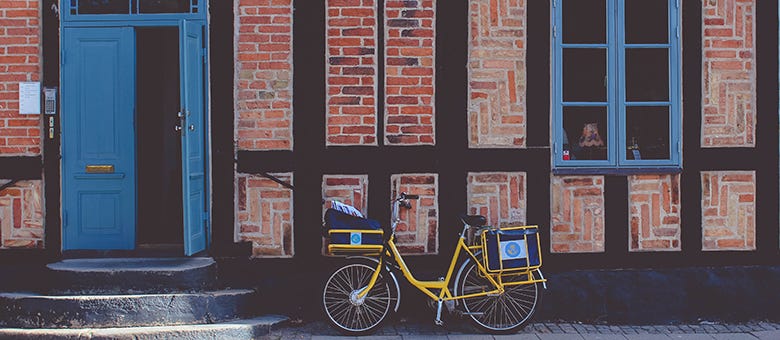 Gelbes Lastenfahrrad steht vor einem Altbau mit Fachwerkfassade; links eine geöffnete blaue Haustür, rechts ein blau gerahmtes Fenster.