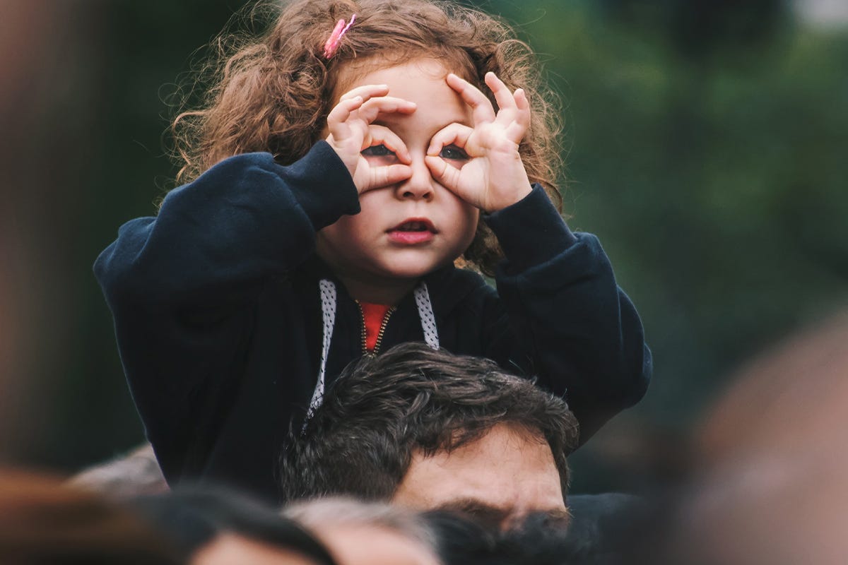 Kleines Mädchen sitzt auf den Schultern ihres Vaters und guckt durch ihre als Brille geformten Finger
